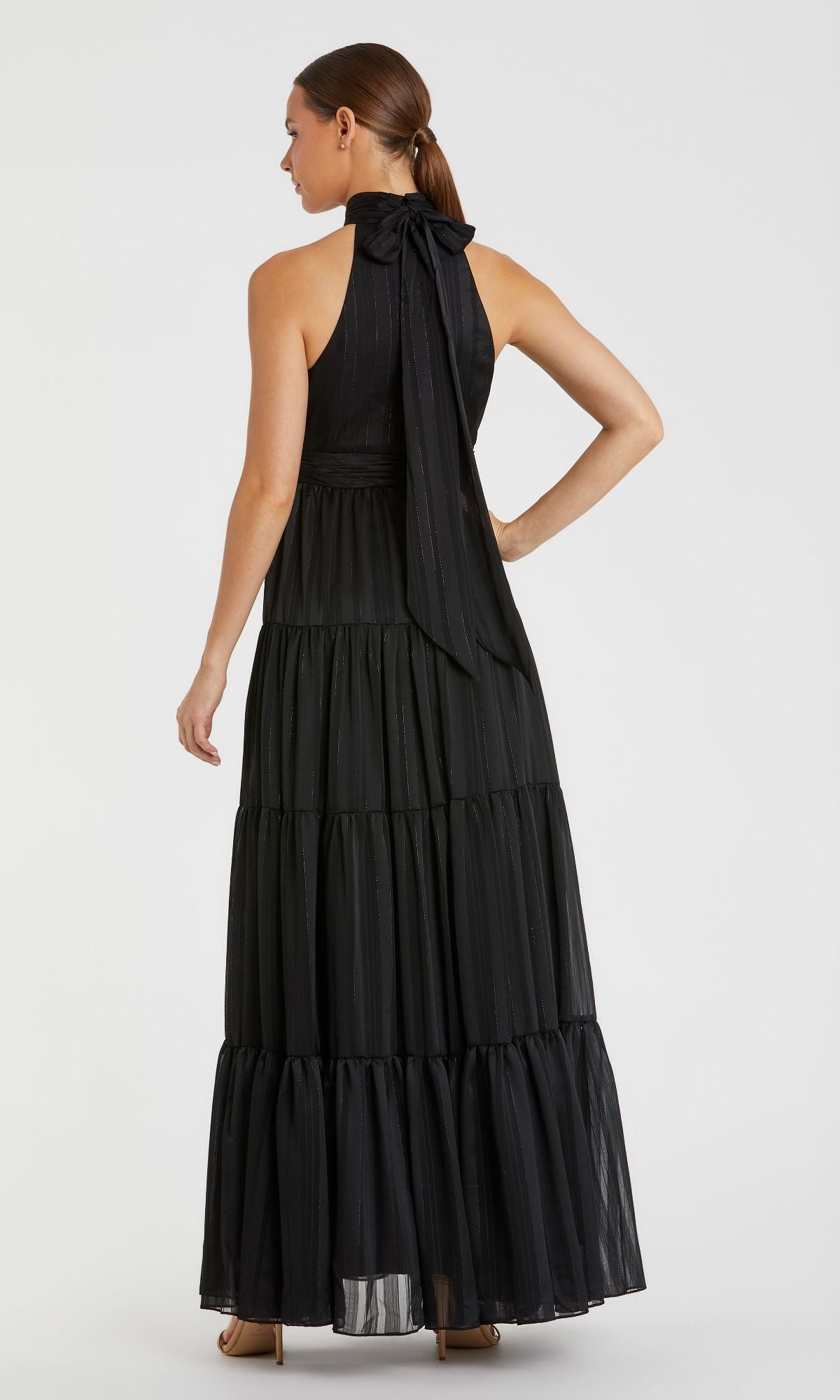 A woman in the Mac Duggal 49515 Long Black Tiered Formal Dress, featuring a high neck and large bow detail, pairs it with nude heels against a plain light background—a chic take on classic elegance.